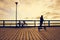 Silhouette of girl with kick scooter and bicycle on sea bridge at sunset.