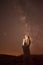 Silhouette of hooded person at night holding illuminated cross with milky way
