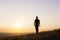 A silhouette of a lone man with a rucksack walking towards the camera on a hill with the sun setting across the landscape on a sum