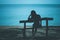 Silhouette of lonely woman sitting on wooden bench on the beach and looking to blue sea.