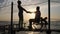 Silhouette of Lovers on wheelchair standing to quay on background of water and sky in sunset