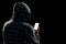 Silhouette of a man in a hood on a black background, his face is not visible, the hacker is holding the phone in his hands. The