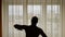 Silhouette of a man performing exercise indoors. In the background city street behind window curtains. Men warming up do