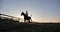 Silhouette of a rider gallops across the farm at sunset.