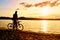 Silhouette of sportsman holding bicycle on lake bech, colorful sunset cloudy sky and reflection in wavy water level