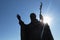 Silhouette of statue of Pope John Paul The Second with sun behind his left hand with cross, clear blue skies in background.