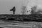 A silhouette of a surfer walking across rocks as a wave crashes against the rock in the distance