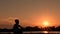 Silhouette of a teenager against a background of sunrise. The guy meets the dawn sitting on the river bank. Backlighting. Copy spa