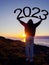 Silhouette of a teenager girl holding sign 2023 above her head. Sunrise sun in the background. Welcoming new year concept. Warm