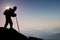 Silhouette of tourist with backpack and poles in hands stand on rocky view point and watching into morning landscape below.