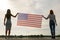 Silhouette of two young friends women holding USA national flag in their hands standing together outdoors. Patriotic girls