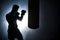 Silhouette of an unrecognizable boxer man sparring with a punching bag in a dark gym. Male athlete working out in