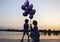 Silhouette of a woman and a child holding a bunch of balloons in their hands, stand by the lake at dusk at sunset