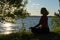 Silhouette of a woman sitting in a lotus position on the shore of a lake in summer. Meditation. Sports in old age