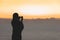 Silhouette of woman with wet hair wrapped in a blanket after swimming. Female taking picture on mobile phone standing on the beach