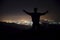 Silhouette of a young man with raised-up arms, standing on the top of the mountain under night sky with stars and near city.