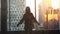 Silhouette of young woman stands by the big window stretches arms and looking out her apartment on the city buildings