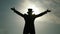 Silhouetted against the bright sky a figure stands with arms outstretched back facing the camera. Dressed in a top hat