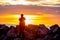 Silhouetted figure standing atop a rocky outcropping at the edge of an orange-hued sunset