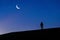 Silhouetted person walks by the hill with a camera and looking at young moon at Morning Twilights