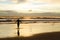 A silhouetted woman with open arms near the sea on a beach with the sun rising and the sunrays reflecting in the sea water.