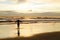 A silhouetted woman with open arms near the sea on a beach with the sun rising and the sunrays reflecting in the sea water.