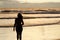 A silhouetted woman walking near the sea on a beach with the sun rising and the sunrays reflecting in the sea water.