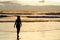 A silhouetted woman walking near the sea on a beach with the sun rising and the sunrays reflecting in the sea water.