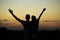 Silhouettes of father and daughter on his shoulders with hands up having fun, against sunset sky.