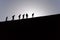 Silhouettes & shadows of six people hiking up a tall sand dune in Sossusvlei, Namib Desert, Namibia shortly after sunrise.