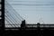 The silhouettes of two people crossing a suspension bridge on foot. Photo during the day.