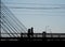 The silhouettes of two people crossing a suspension bridge on foot. Photo during the day.