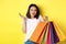 Silly asian girl showing shopping bags and shrugging, shop in stores with discounts, standing over yellow background