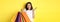 Silly asian girl showing shopping bags and shrugging, shop in stores with discounts, standing over yellow background