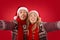 Silly young couple in Santa hats showing tongues while taking Christmas selfie on red background