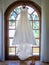 Simple wedding dress hanging from a big window with a garden under the sunlight on the background