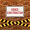 Simple white sign with text `Under Construction` hanging on a red brick wall with warning tapes. Brickwork background.