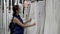 A single adult woman in a medical mask examines and evaluates white curtains in an empty shop floor before buying