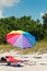 Single colorful beach umbrella a beach lounge and beach shoes on a sandy, isolated beach