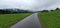Single road between green fields with very cloudy, mysterious hanging clouds over the far away mountains