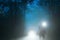 A sinister hooded figure standing in front of a car headlights on a spooky forest road on a misty evening