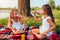 Sisters drinking juice while having picnic by summer river at sunset. Kids eating cake and relaxing with family