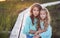 Sisters relaxing outdoors while sitting on a wooden path in a field at sunset.