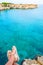 Sitting on the edge of a cliff with dangling legs in travel sandals looking on crystal clear water on Torre Sant Andrea beach with