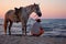 Sitting man with standing horse on the beach by the sea at sunset.