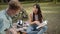 Sitting on the plaid in a public park, a young asian girl with long dark hair is reading a book for her disabled friend