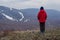 A six-year-old boy in a red warm jacket and sneakers stands with his back to the camera and looks at the high mountains in the fog