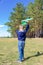 A six-year-old preschooler boy in a blue jacket launches a toy plane in a field against a blue sky with clouds on a summer day