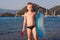 A six year old tanned boy stands holding a swimming circle and a sand shovel by the sea