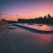 A skatepark equipped with various ramps and obstacles is aglow with the warm hues of a setting sun on the horizon.